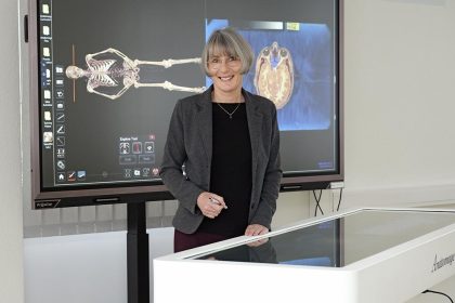 Freundlich lächelnde Dozentin steht vor einem großen Bildschirm, auf dem gerade Lehrmaterial zum menschlichen Skelett eingeblendet ist. Der große Bildschirm ist mit einem modernen Gerät, dem Anatomage, verbunden.