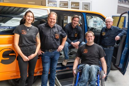 5 Menschen, 4 Männer - Davon 1 junger Mann im Rollstuhl sitzend und auf der linken Seite eine junge Frau, befinden sich freundlich lächelnd vor einem seitlich geöffneten Fahrzeug