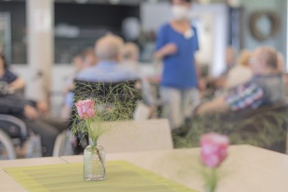 Im Vorderund ein einladend hergerichteter Tisch mit 2 Rosen, welche jeweils in einem Glas stehen, im Bouquet des Hintergrunds sind sitzende ältere Menschen zu erkennen, in deren Mitte eine stehende Mitarbeiterin mit Mundschutzmaske.