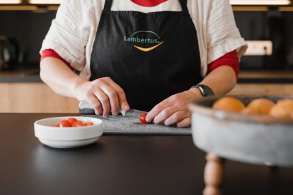 Ein Mensch, gekleidet mit einer Schürze, auf der das Lambertus-Logo ist, schneidet kleine Tomaten, um eine leckere Mahlzeit zuzubereiten.