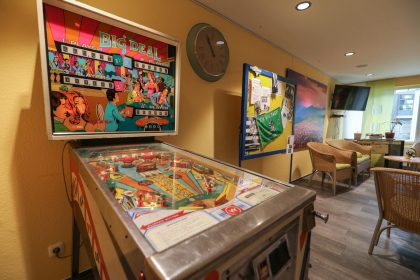 Ein klassischer Flipper steht in einem Aufenthaltsraum des Hermann-Josef-Altenheim. Im Hintergrund ein Clipboard, mehrere Sitzmöglichkeiten und ein modernes TV-Gerät.