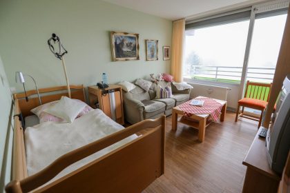 Impression eines Zimmers, Wohnraum im Hermann-Josef-Altenheim mit Bett, Nachttisch, Couch, Tisch, Stuhl und TV sowie Außensicht über das Fenster.