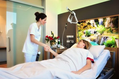 Junge Klinikmitarbeitern, Ärztin gibt freundlich lächelnd einer im Bett liegenden Patientin die Hand