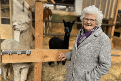 Seniorin steht lächelnd vor einem Holzzaun eines Alpaka oder Lama-Geheges